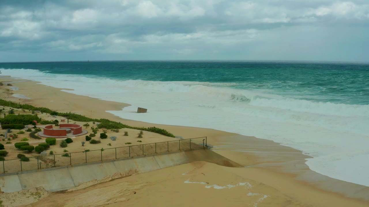 At 0:03, Dreams Resort Los Cabos surf covers much of the midground sand, with the fence and landscaped beach edge visible below and no visible people.