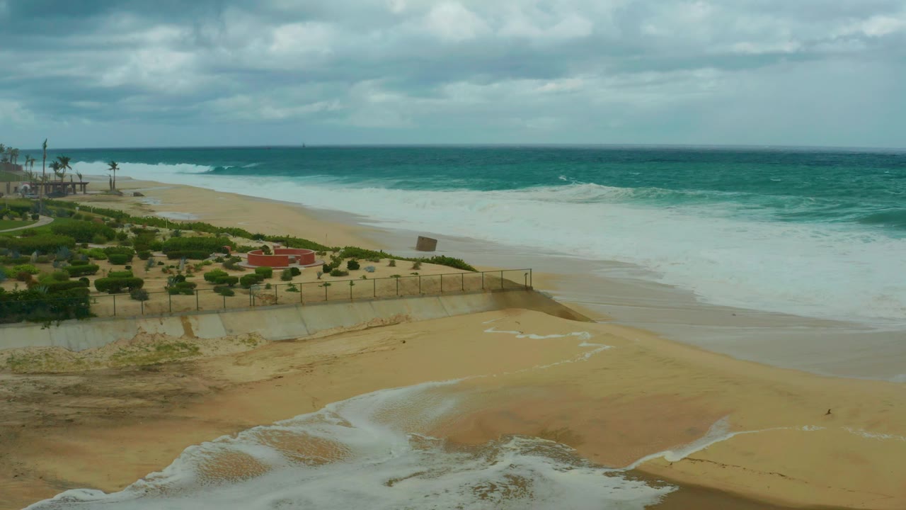 Opening frame over Dreams Resort Los Cabos beach, showing foamy surf, a runoff stream on wet sand, dune vegetation, a fence, palms at left, and no visible people.