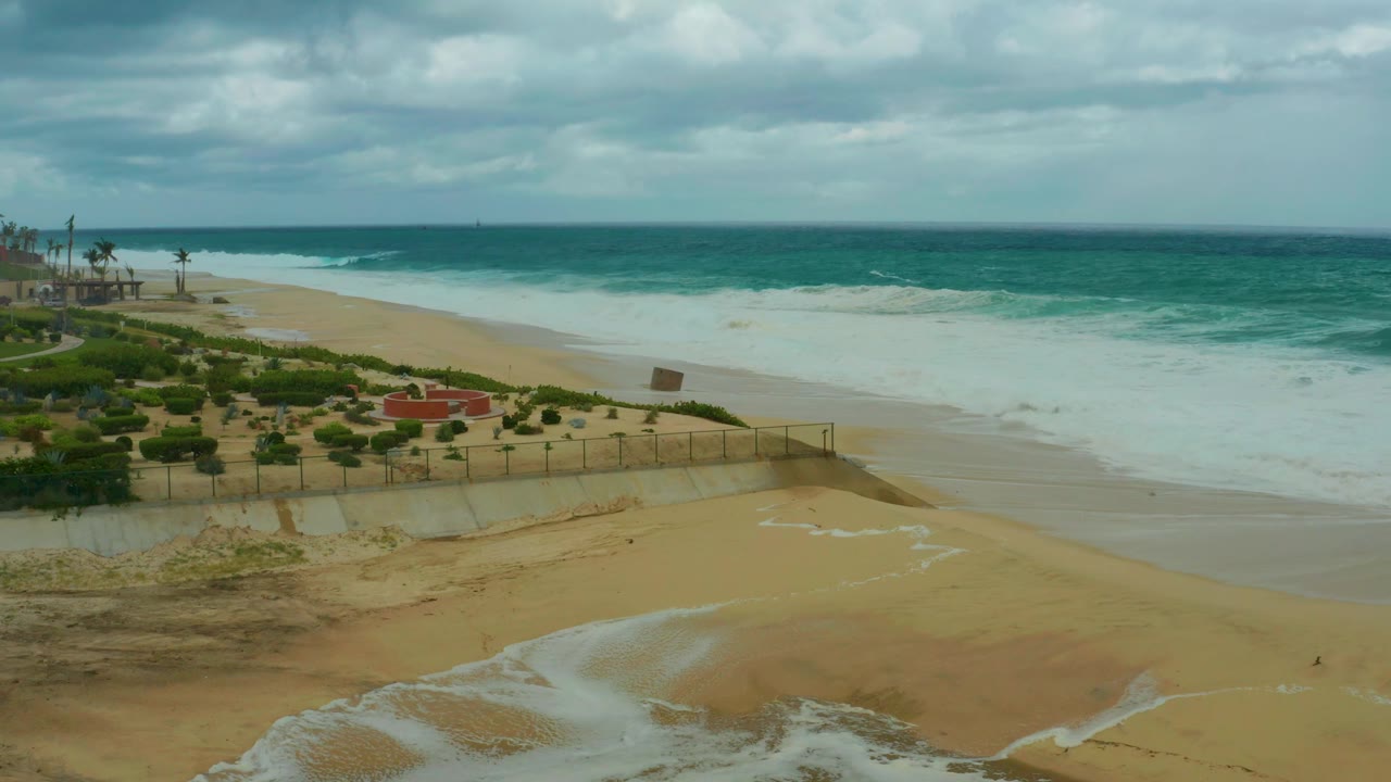 Elevated view at Dreams Resort Los Cabos with a broad sheet of foam along the beach, a thin water channel in the foreground sand, fenced landscaping, and no visible people.