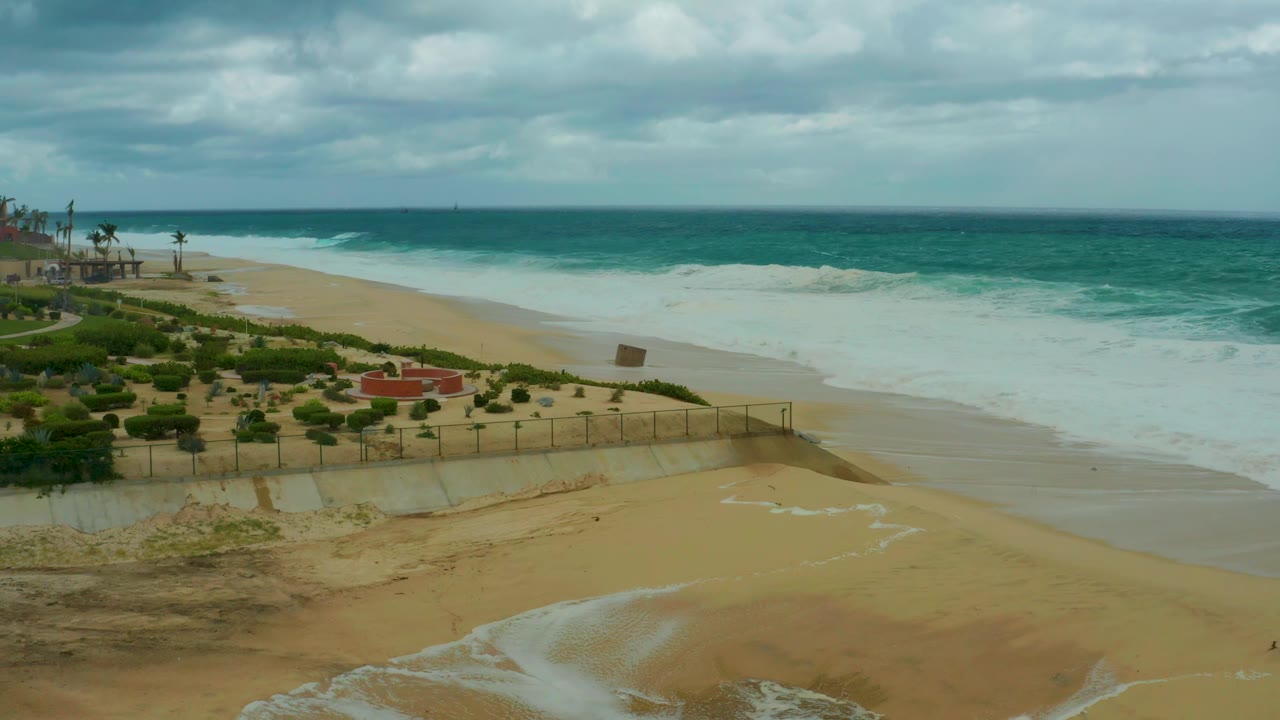 Cloudy ocean beach scene with waves, wet sand, dune fence, and palms.