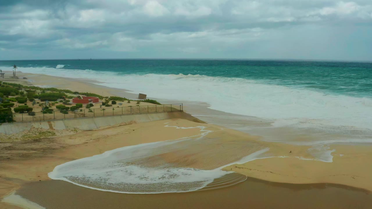 At 0:01, the wave foam forms a broad curved sheet over the lower beach at Dreams Resort Los Cabos, with ocean horizon, clouds, fencing, vegetation, and no visible people.