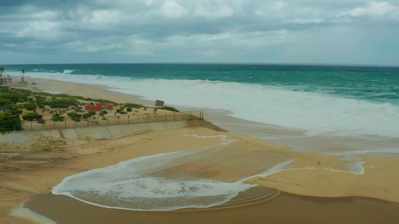 At Dreams Resort Los Cabos, surf foam lies in a rounded pool across the sand while blue-green water and a cloudy horizon fill the upper frame, with no visible people.