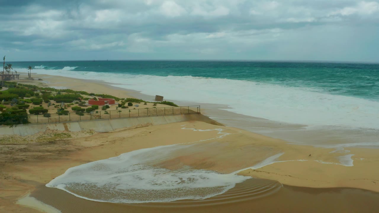At 0:03, the foamy wash thins over the foreground sand at Dreams Resort Los Cabos while incoming surf and low clouds remain visible beyond the fence, with no visible people.