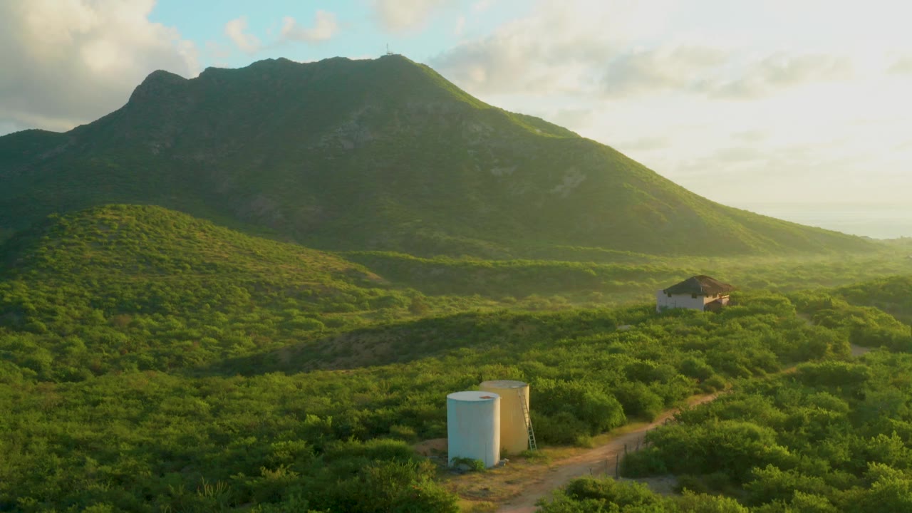 At 0:00 in Cabo Pulmo, Baja California Sur, green vegetation surrounds a dirt road and white tanks below a small building and broad mountain, with no visible people.