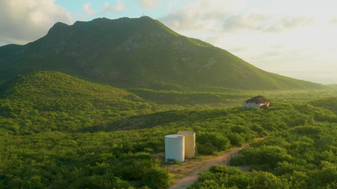 At 0:02 in Cabo Pulmo, warm light catches the green valley as the white tanks sit near the lower frame and the small building remains on the hillside, with no visible people.