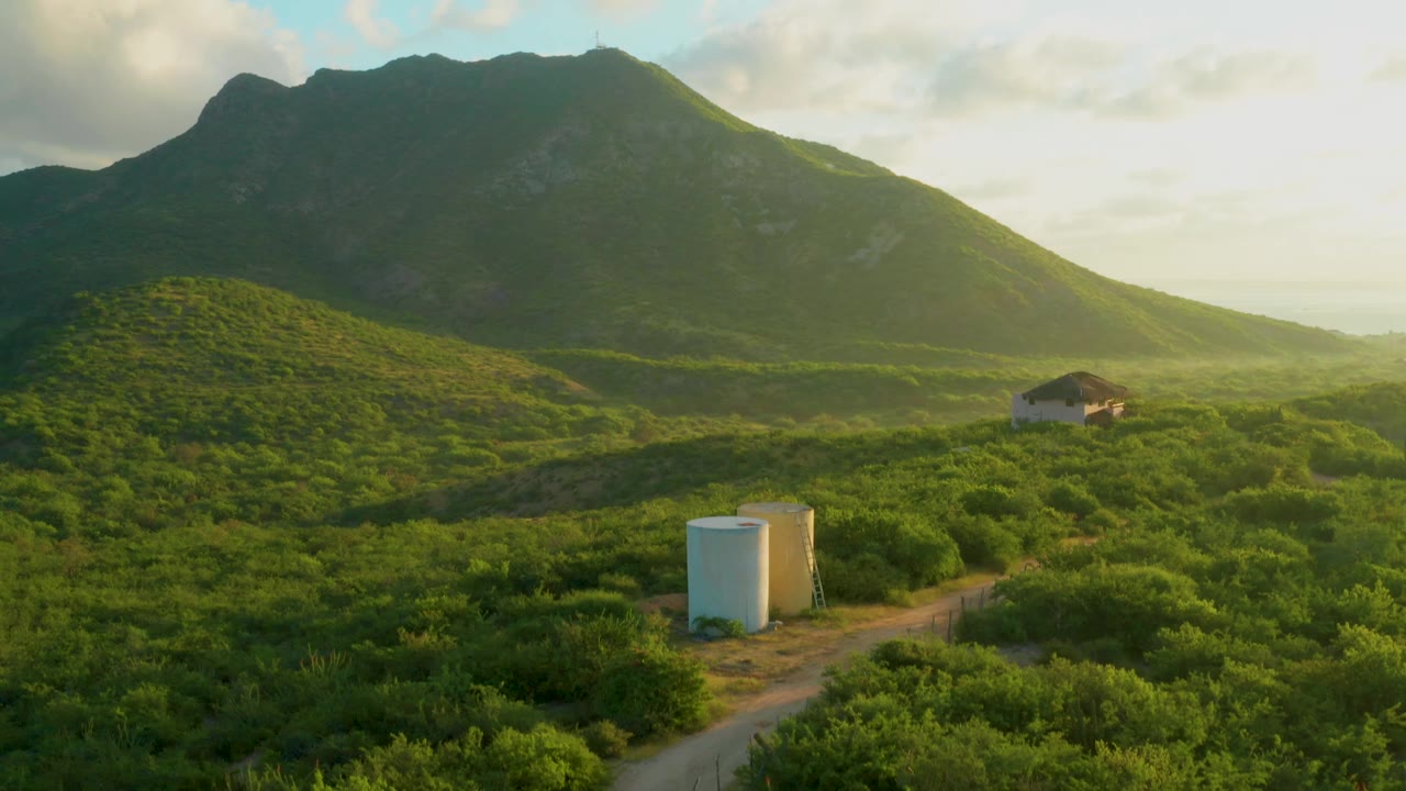 Mountain valley with dirt road, two white tanks, green shrubs, and a small white building.