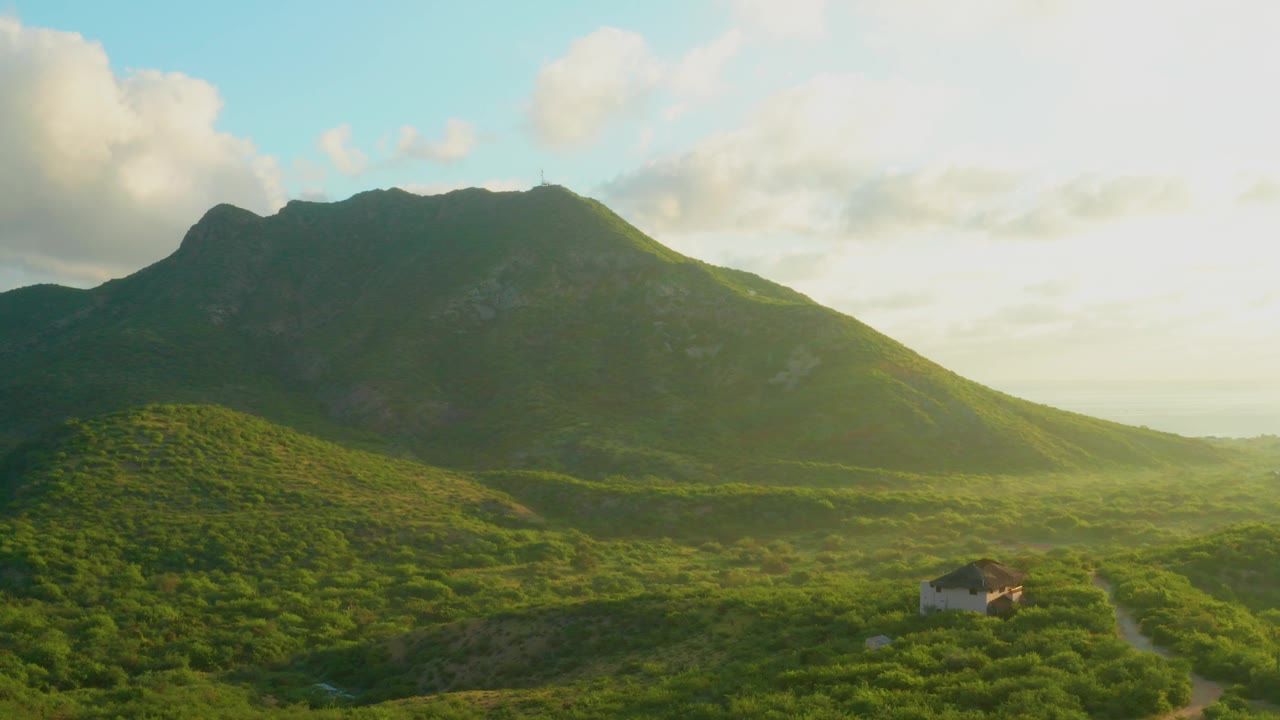 Wide aerial view of a green Cabo Pulmo mountain slope with a small house low in frame, bright clouds above, and no visible people.