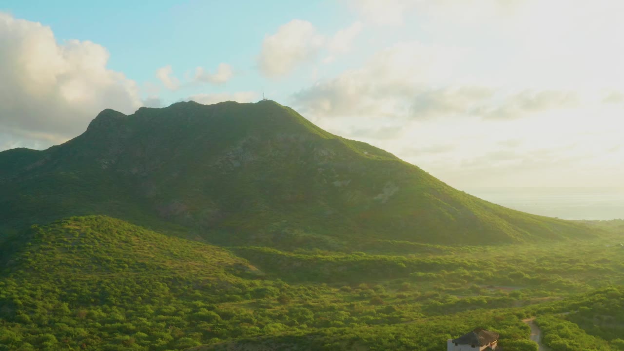 Aerial view of a green valley below a dark mountain ridge and bright clouded sky.