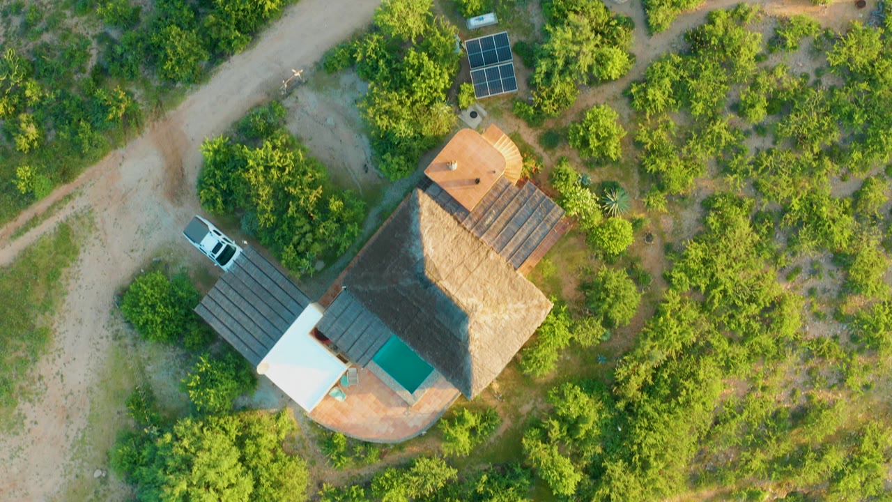Overhead frame in Cabo Pulmo shows a thatched-roof house bordered by green trees, pale dirt roads, solar panels, and a white car, with no visible people.