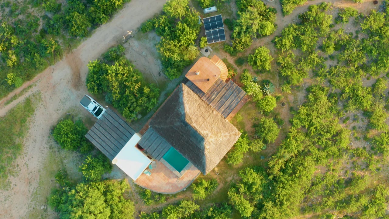 Aerial view of the thatched roof house with solar panels, a white car, and surrounding greenery.