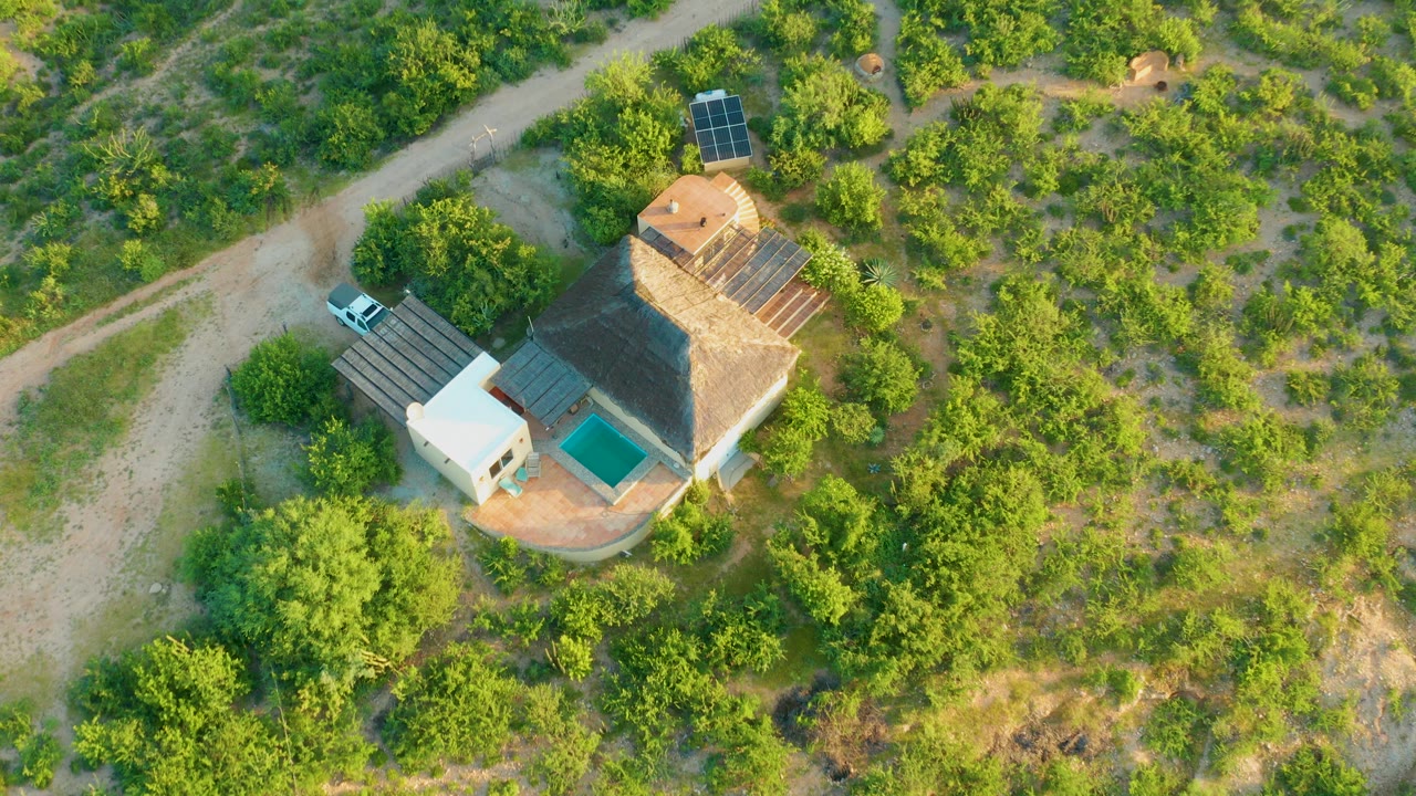 Overhead drone frame in Cabo Pulmo, Baja California Sur, showing a thatched-roof house, solar panels, dirt road, parked white car, and surrounding green brush with no visible people.