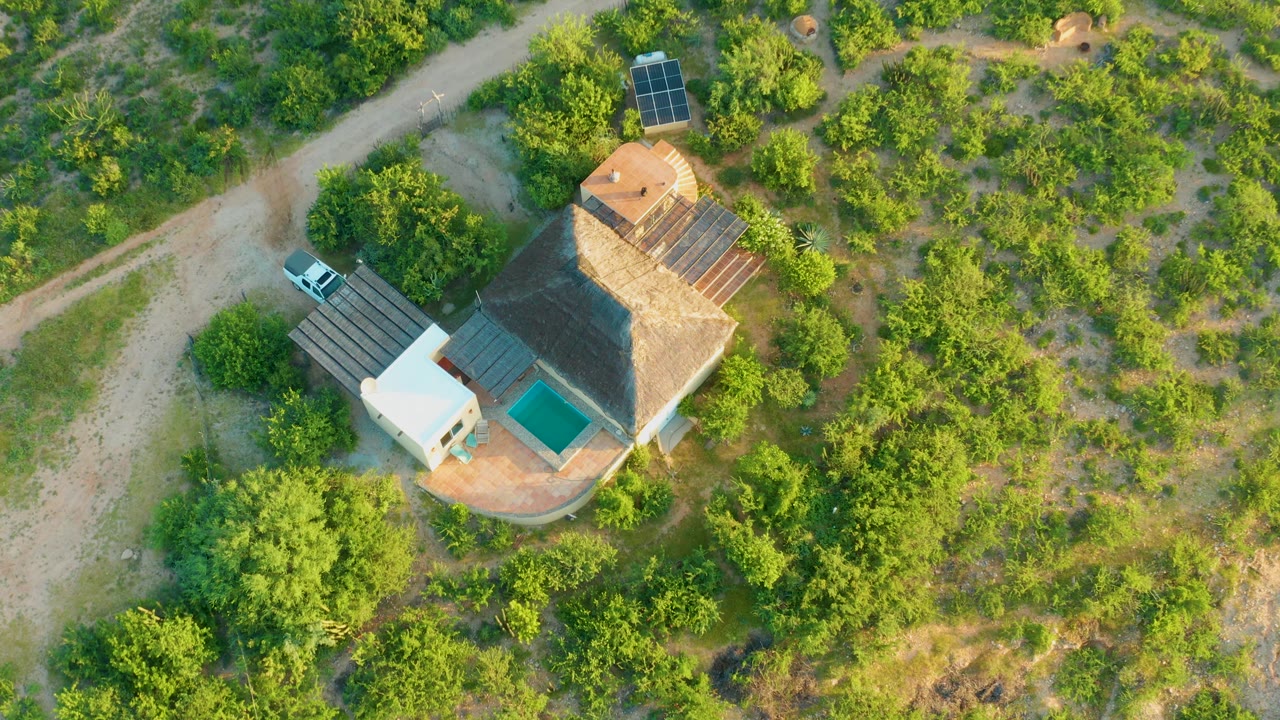 Overhead Cabo Pulmo frame showing the thatched house near a pale dirt road, a parked white car, solar panels above the property, and dense green vegetation with no visible people.