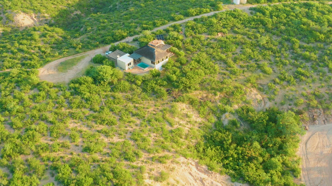 Wide aerial view in Cabo Pulmo, Baja California Sur, showing a small house complex on a green hillside with a winding dirt road and no visible people.