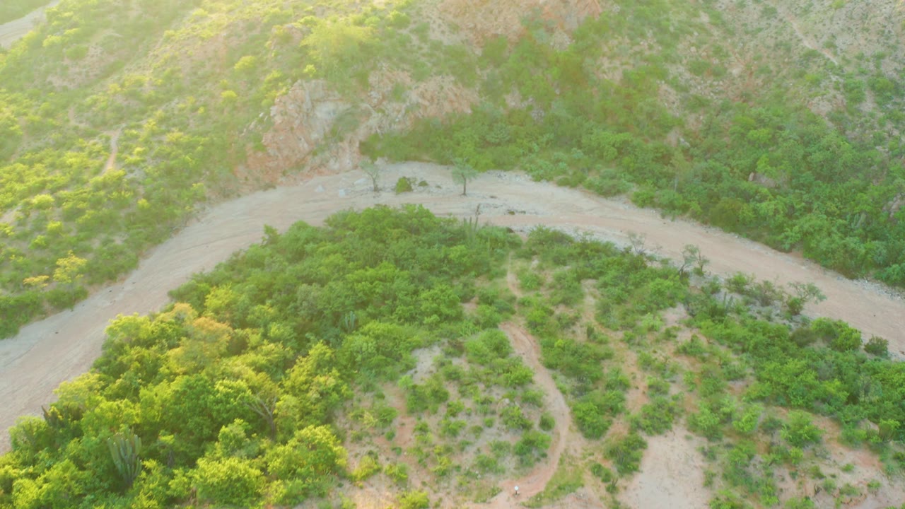 The drone view looks down on the Cabo Pulmo dirt road as it arcs across a green hillside, with a small vehicle on the road and no visible people.