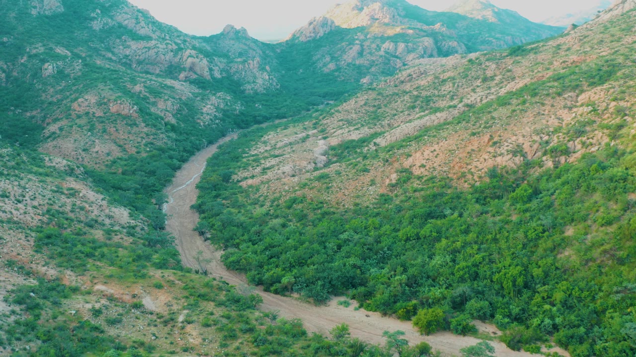 Cabo Pulmo aerial view with the river running from the lower frame into the green valley, flanked by pale rocky slopes and distant ridges, with no visible people.