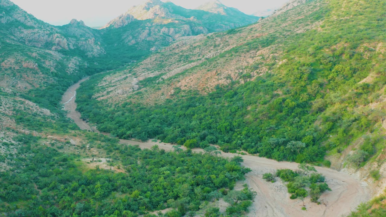Aerial view in Cabo Pulmo, Baja California Sur, showing a pale winding riverbed below green valley vegetation and rocky mountain slopes, with no visible people.
