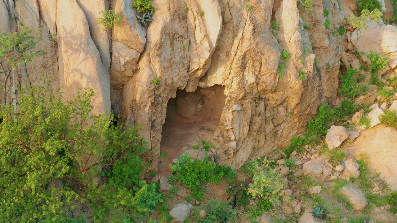 Tan rock wall, cave opening, shrubs, and scattered stones.