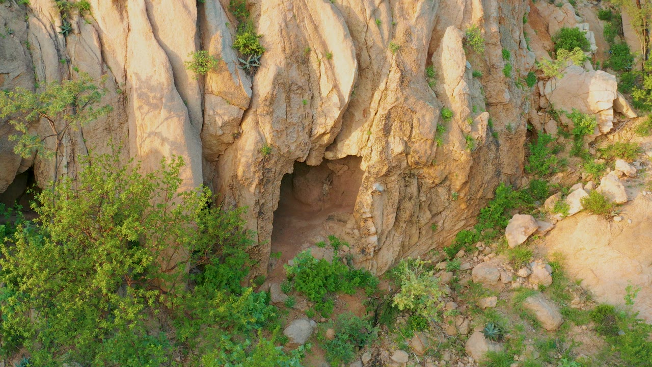 High angled view of a tan rocky cave opening in Cabo Pulmo with green shrubs, scattered boulders, sandy ground, and no visible people at the start of the clip.