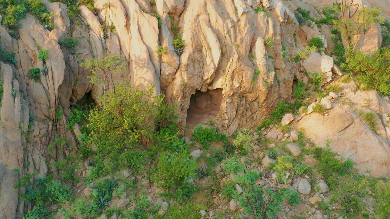Cabo Pulmo aerial view with the cave opening low on the left, vertical tan rock ridges above it, dense green shrubs in the center, and scattered boulders on dry ground, with no visible people.