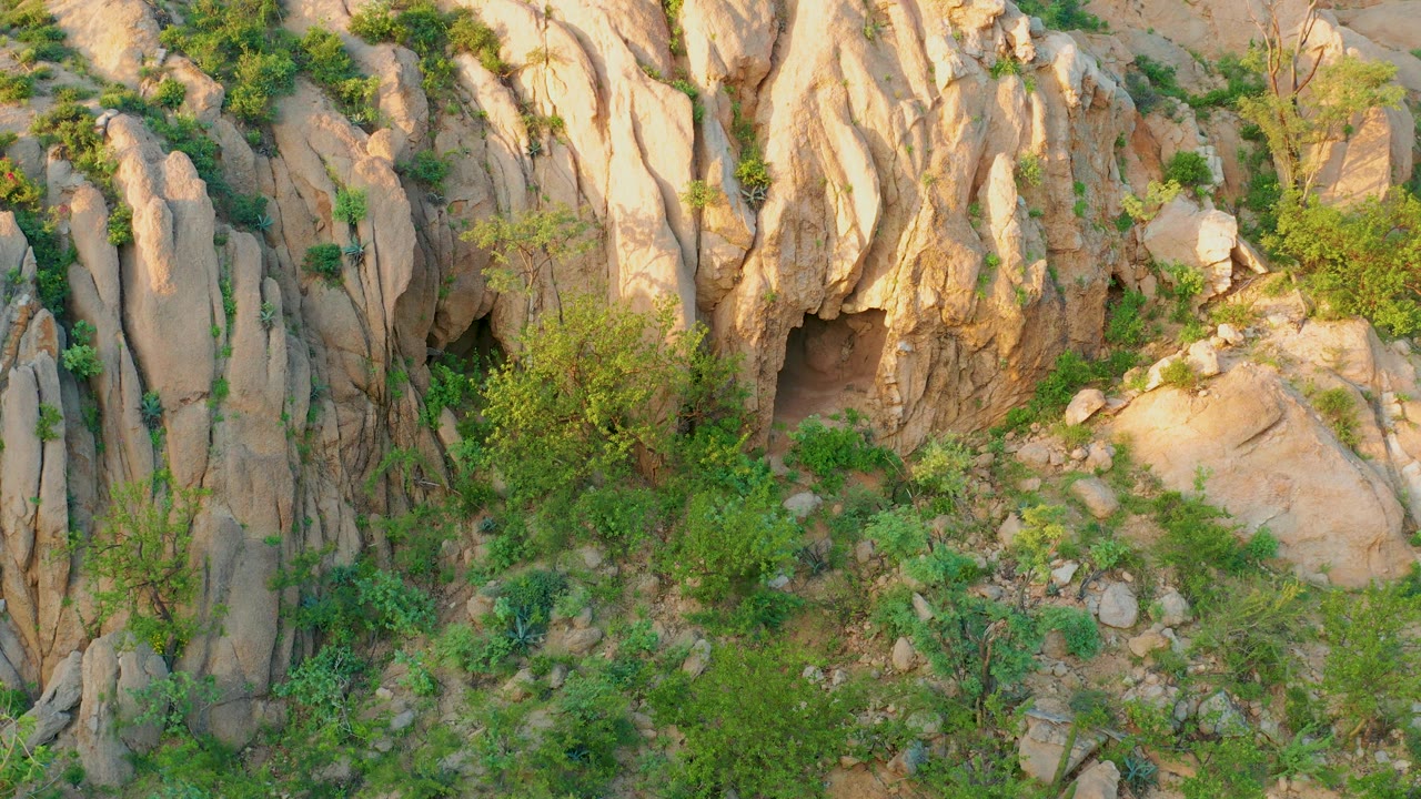 Cabo Pulmo rock formation with a small dark recess near the left side, sunlit ledges, green brush below, and no visible people.