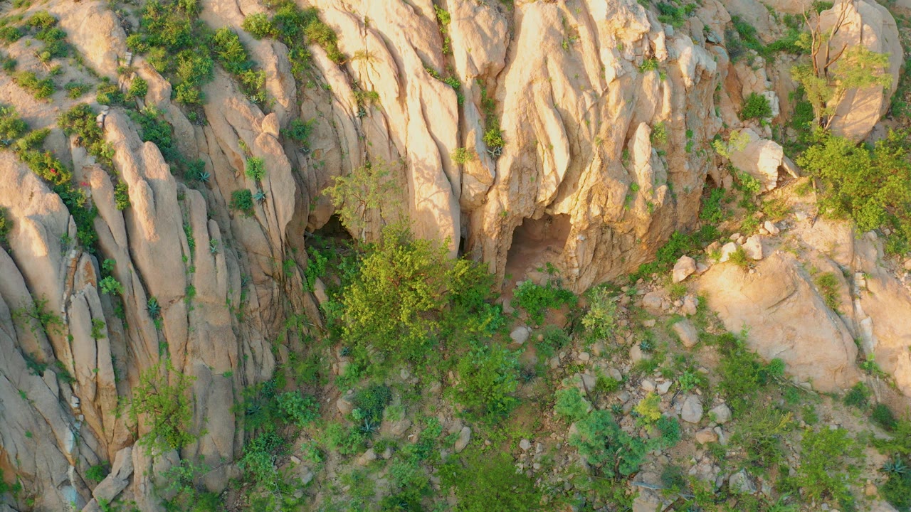 Mid-clip Cabo Pulmo frame showing the dark cave entrance near the lower-left rock face, tan carved-looking stone surfaces, and scrubby green plants across the slope, no visible people.