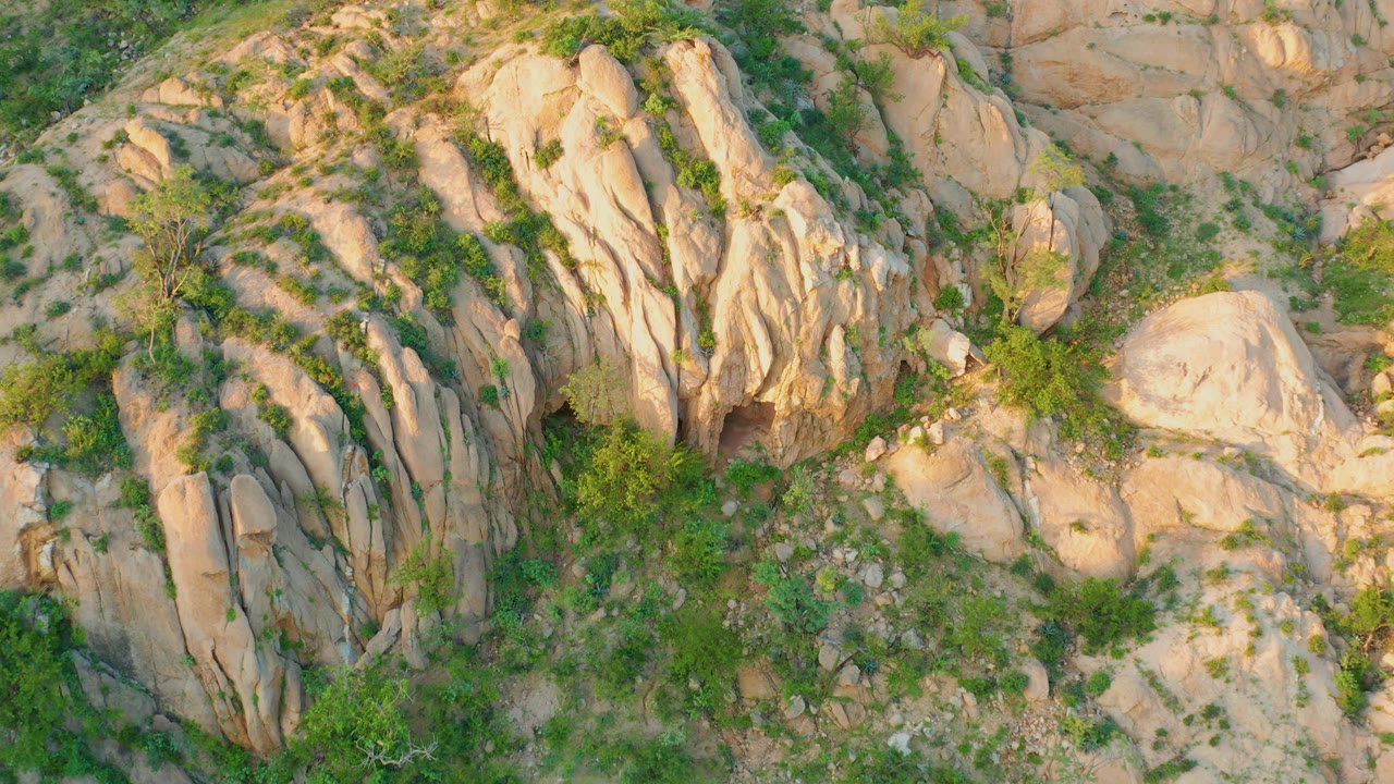 Aerial frame over Cabo Pulmo terrain with ribbed tan rock faces, small green plants, exposed sandy patches, and no visible people.