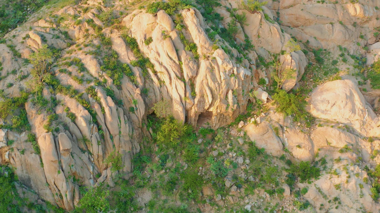 Downward aerial view in Cabo Pulmo showing pale rock slabs, a large rounded boulder at the side, green shrubs in crevices, and no visible people.