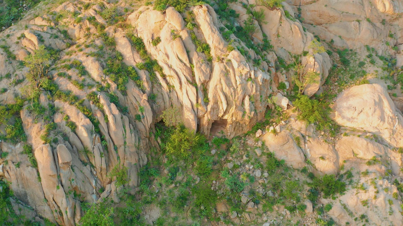 Aerial view of the Cabo Pulmo hillside with warm light on tan stone, sparse green brush between rock formations, sandy tracks, and no visible people.