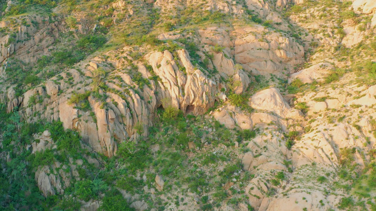 At 0:03, Cabo Pulmo's rocky hillside fills the frame with vertical tan stone ridges, green vegetation in cracks, and warm light across the right side with no visible people.