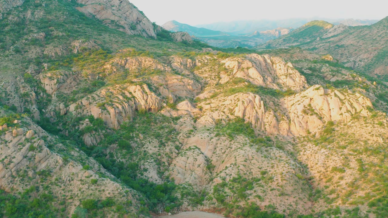 Cabo Pulmo aerial frame where exposed beige rock ridges dominate the midground, green vegetation fills the lower slopes, distant mountains sit under haze, and no visible people appear.