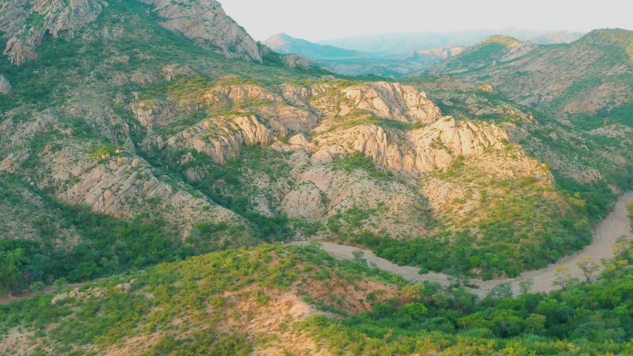 Early frame from Cabo Pulmo, Baja California Sur showing a high aerial view over rocky hills, green valley vegetation, a pale winding road or dry wash, hazy ridges in the distance, and no visible people.