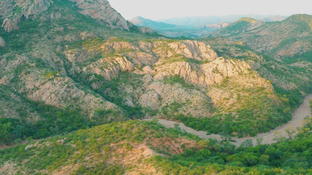 High view across sunlit rock ridges, green brush, and a curving road in a mountain valley.