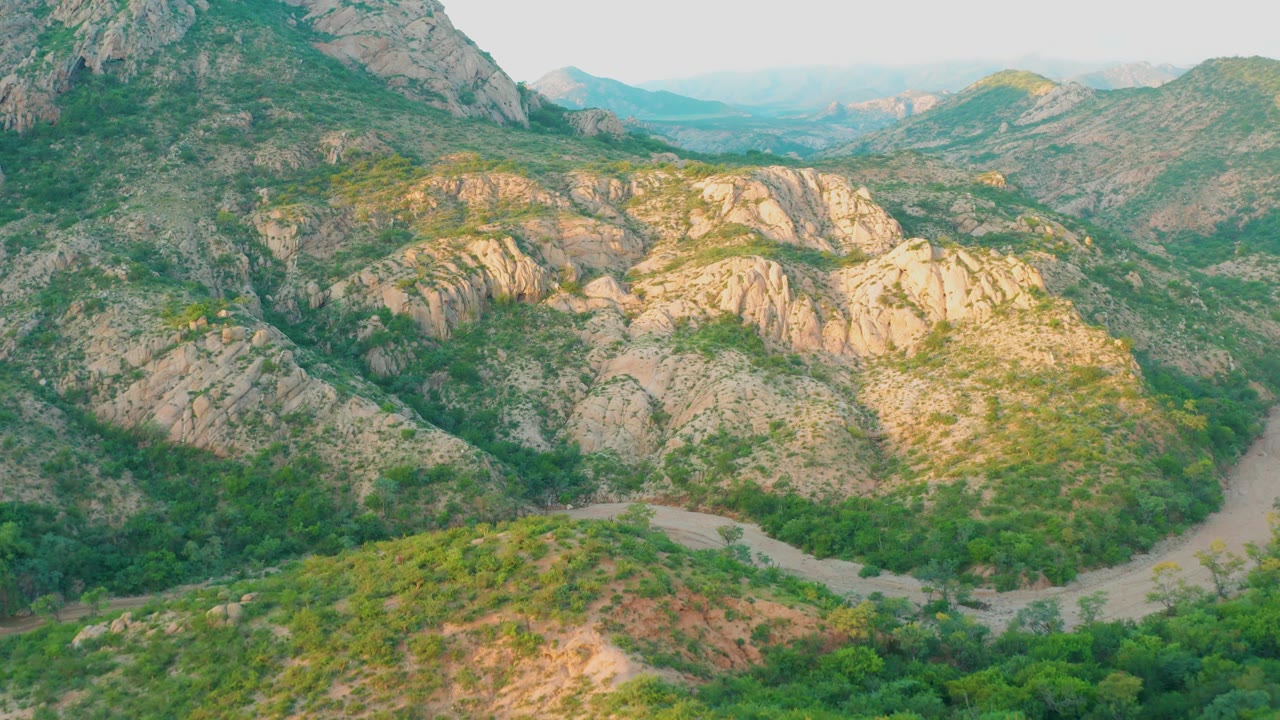 At 0:02, the elevated Cabo Pulmo view shows a rocky mountain slope on the left, a bright rock formation in the center, green trees on the valley floor, a pale route below, and no visible people.