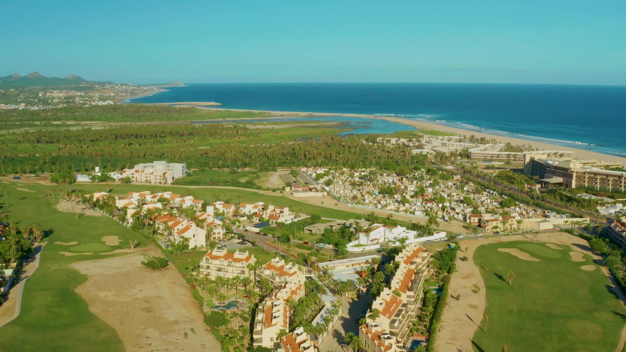 Opening aerial frame over San Jose Del Cabo shows golf fairways and sand traps below, white residential buildings and palms in the middle distance, and blue ocean beyond with no visible people.
