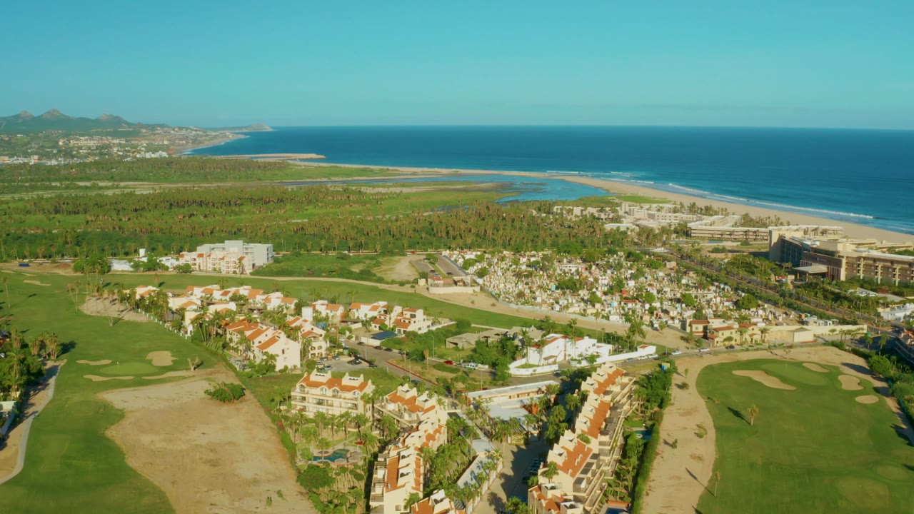 At 0:02, a large resort-style building appears near the right side of the San Jose Del Cabo coastline, with golf fairways and residential blocks below and no visible people.