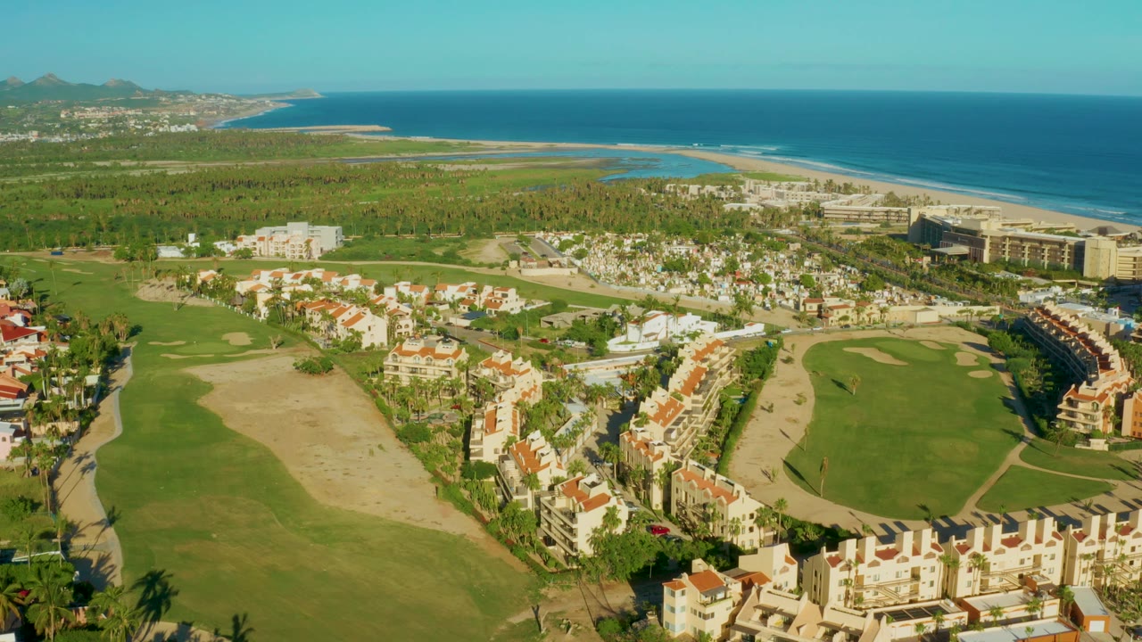 Aerial frame over San Jose Del Cabo showing green golf fairways, sandy hazards, red-roof residences, beach, blue ocean, distant hills, and no visible people.