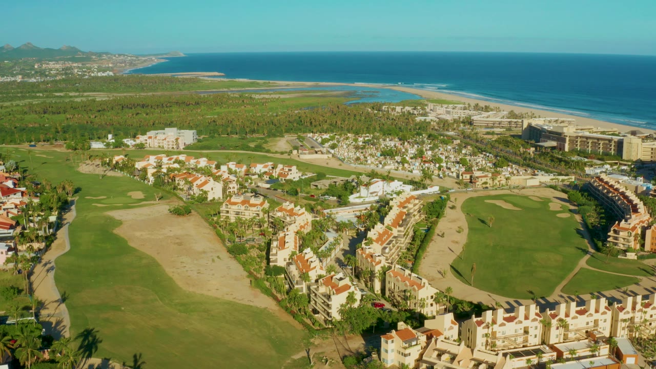 Wide San Jose Del Cabo aerial view with residential buildings around golf greens, palms in the foreground, coastal hotels near the beach, blue ocean, and no visible people.
