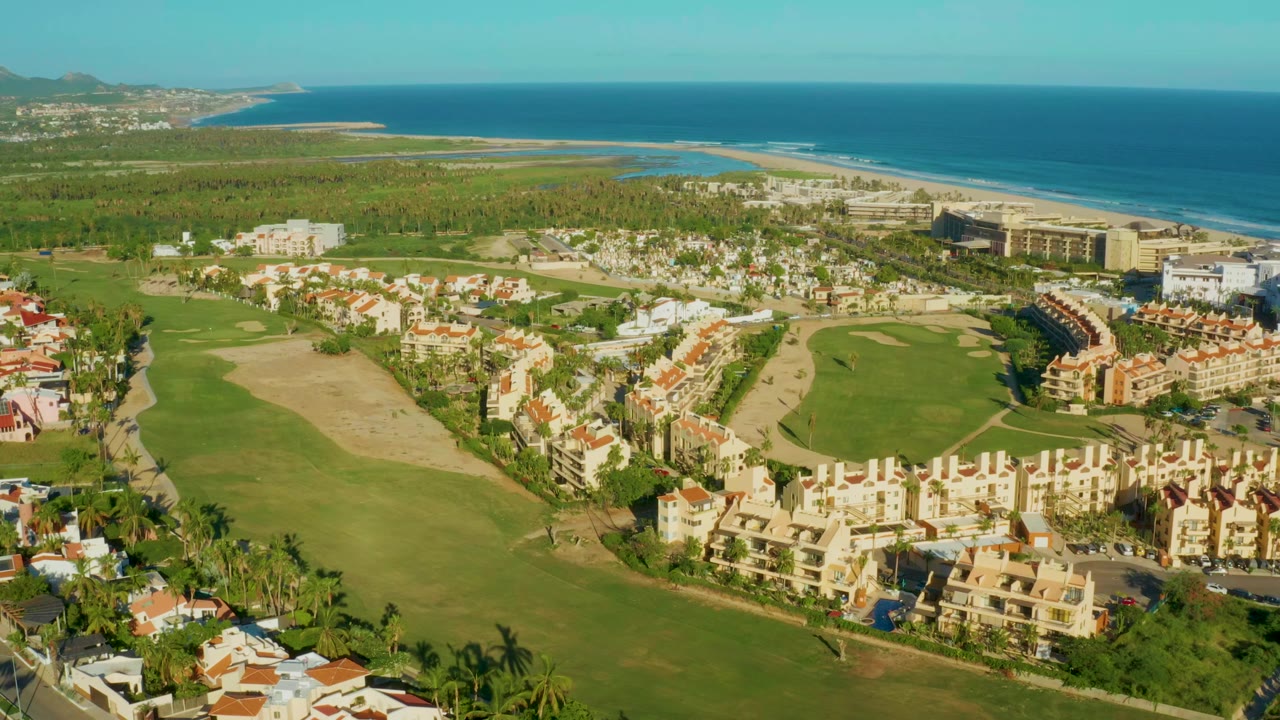 Opening aerial frame over San Jose Del Cabo with golf fairways in front, resort and residential buildings near the shore, blue ocean beyond, and no visible people.