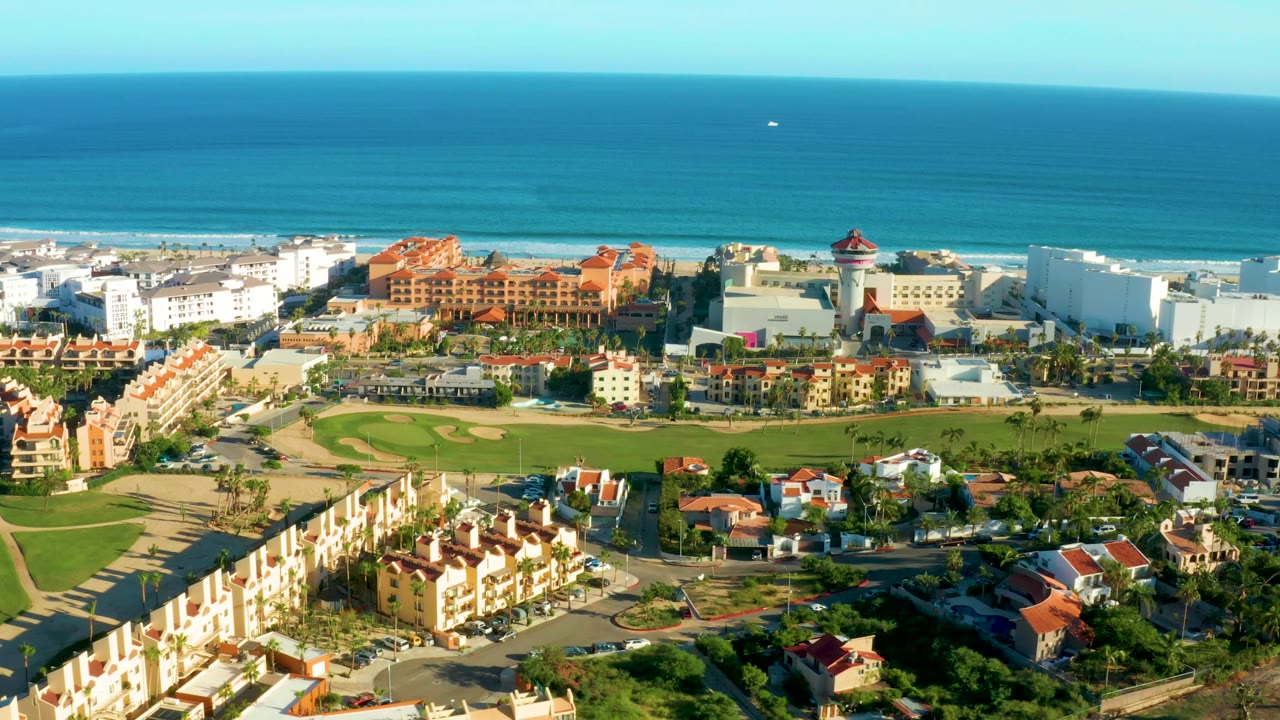 San Jose Del Cabo aerial still showing calm blue water beyond the beach, clustered white buildings, orange-red roofs, a tower-like structure, green grounds, and no visible people.