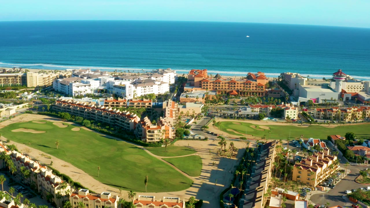 Opening aerial frame over San Jose del Cabo with green golf fairways below, dense residential buildings near the beach, blue ocean beyond, and no visible people.