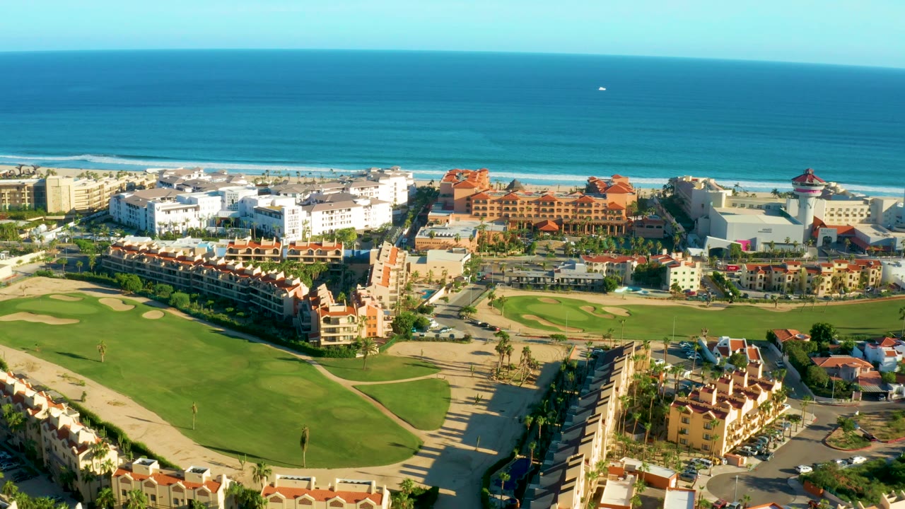 Aerial view in San Jose del Cabo showing the golf course stretching across the foreground, rows of beachside residential buildings, a red-roofed complex, and no visible people.