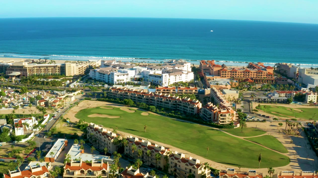San Jose Del Cabo aerial view with the fairway centered lower in frame, resort-style buildings behind it, surf lines along the beach, open ocean, and no visible people.