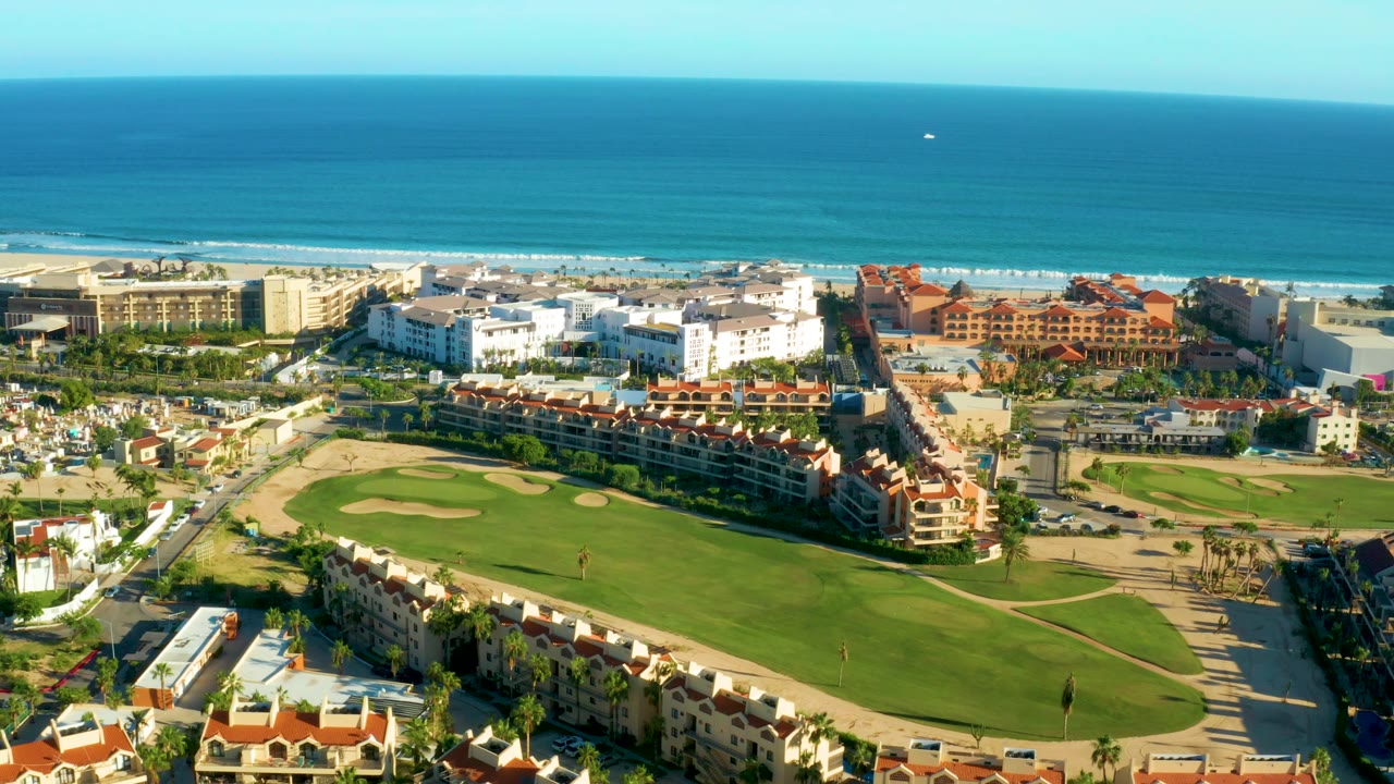 High aerial view in San Jose Del Cabo with red-roofed housing along the lower edge, green golf grounds, bright white buildings, beach strip, blue ocean, and no visible people.
