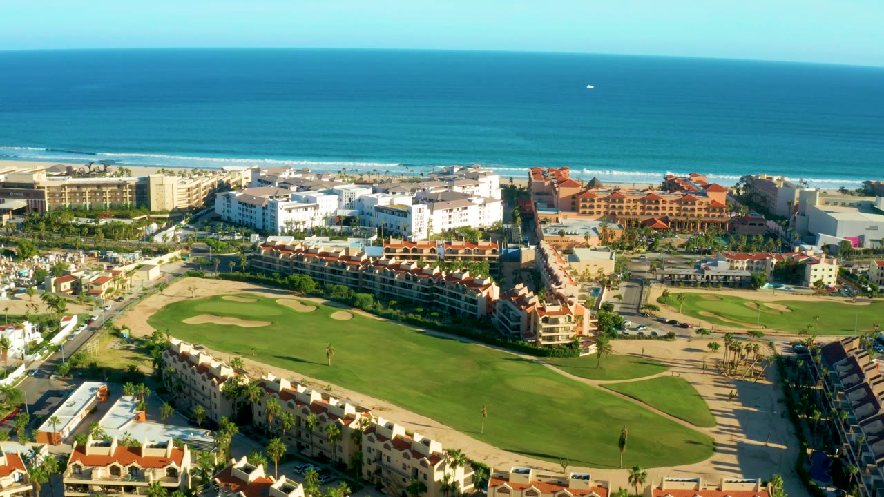 Drone view of a golf course foreground with beach and ocean beyond buildings.