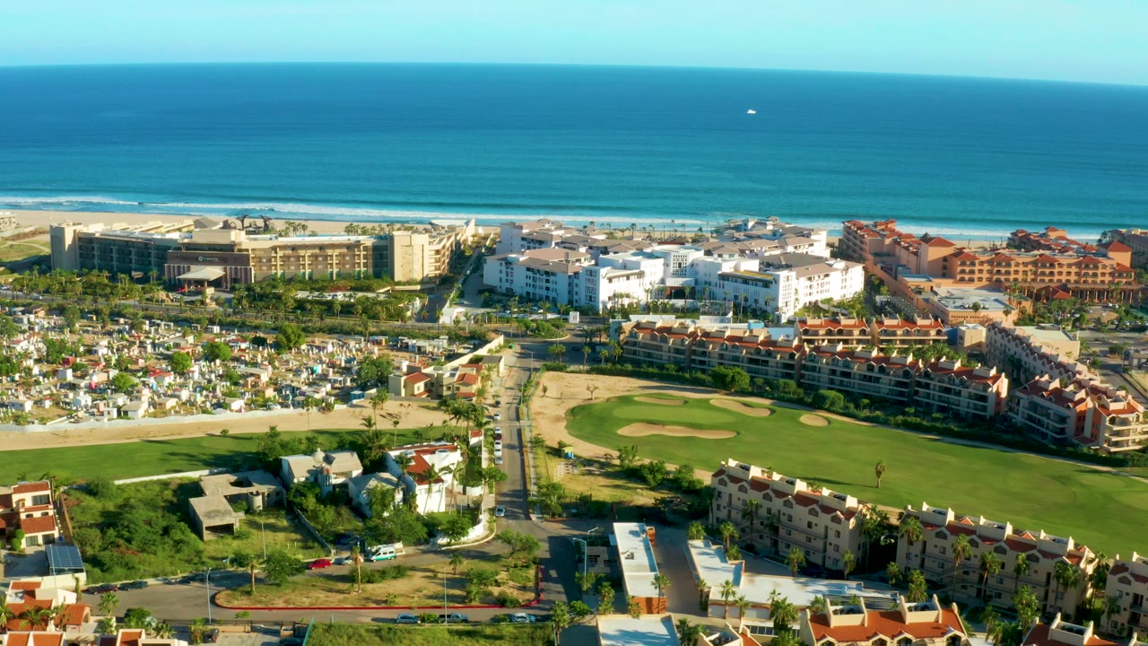 San Jose Del Cabo aerial view showing the fairway crossing the foreground, clustered residential buildings in the center, and the ocean and pale surf bands in the background, with no visible people.