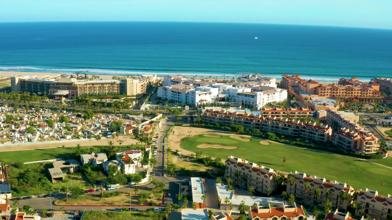 A broad drone view in San Jose Del Cabo shows manicured green golf holes in front of low residential buildings, larger beachfront structures, and deep blue ocean under clear sky, with no visible people.