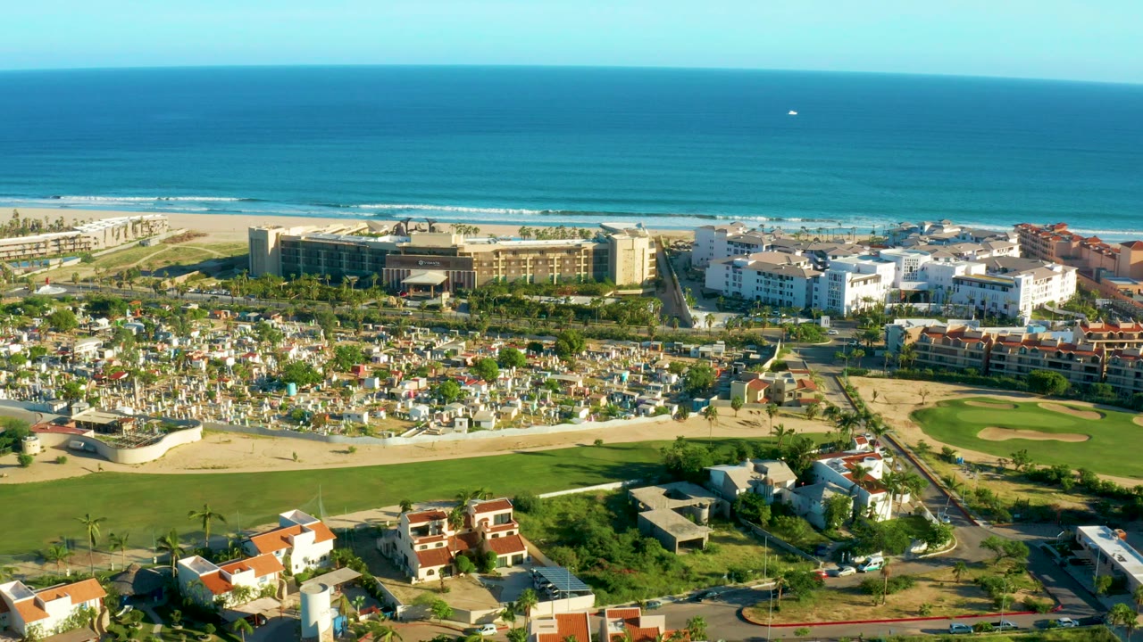 Aerial frame over San Jose Del Cabo showing white hotel buildings, red-roof residences, a green field, sandy beach, and blue ocean beyond, with no visible people.