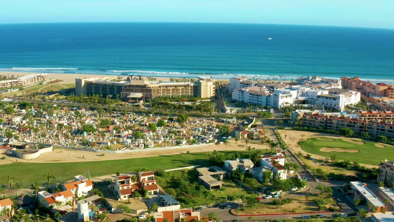 A wide San Jose Del Cabo drone frame shows turquoise water, a pale sand beach, clustered white buildings, red-roof residences, a green field, and foreground roads, with no visible people.