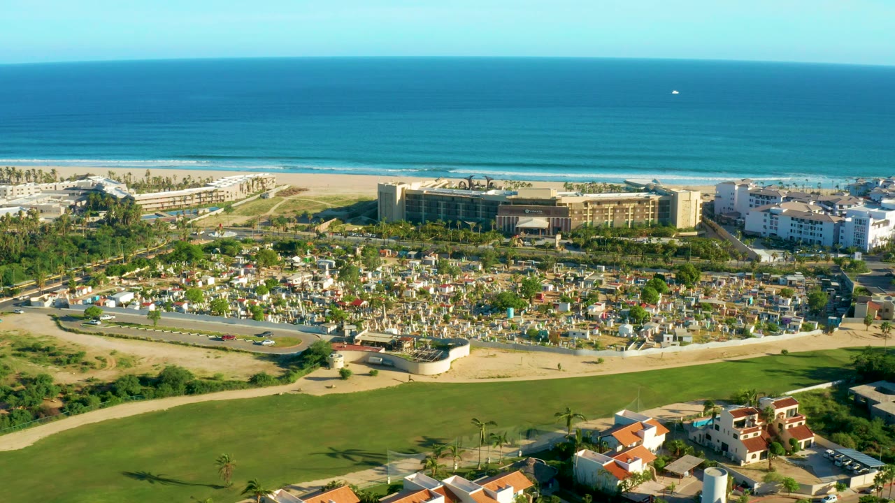Wide aerial frame over San Jose del Cabo showing turquoise ocean, sandy beach, beachfront resort buildings, golf course grass, roads, and red-roofed homes with no visible people.