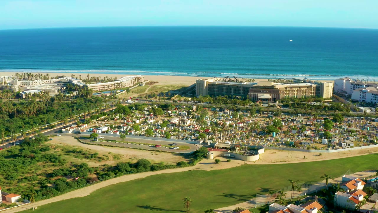 Wide aerial frame of San Jose del Cabo with green foreground, a curved road, clustered coastal buildings, beach, turquoise ocean, and no visible people.